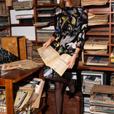 Kobieta w eleganckiej sukience z motywem ptaków, czytająca gazetę w stylowym wnętrzu z półkami pełnymi książek i czasopism.