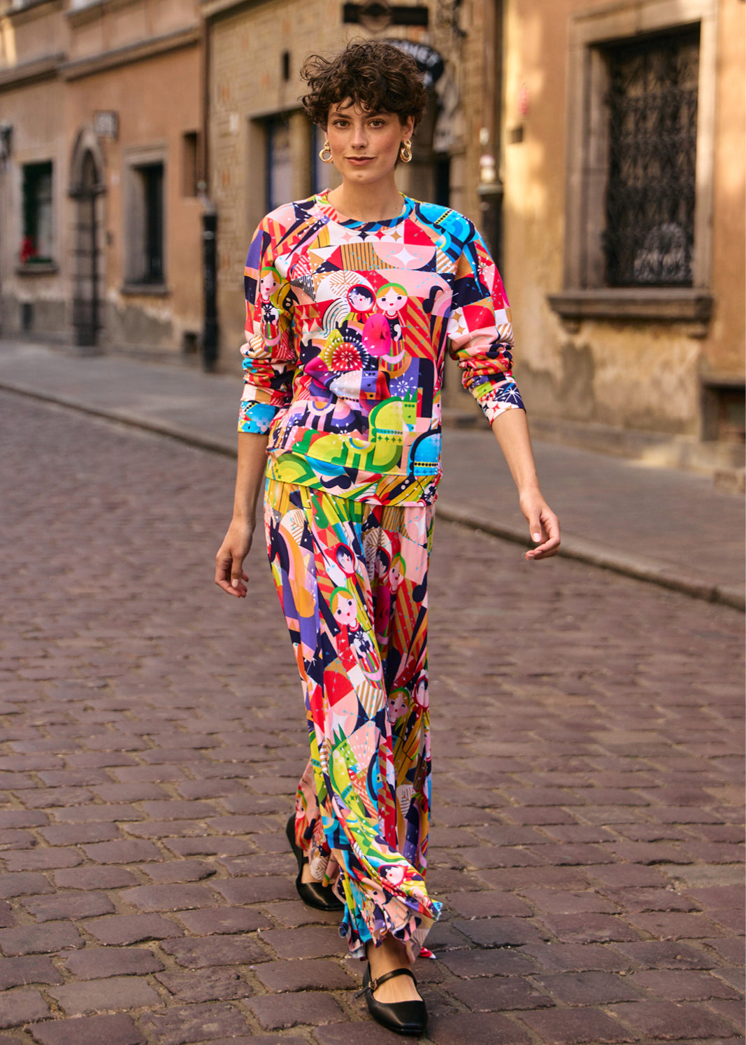 Kobieta w kolorowej, graficznej sukience z motywem lalek, spacerująca po brukowanej ulicy. Moda polska, self-care fashion.